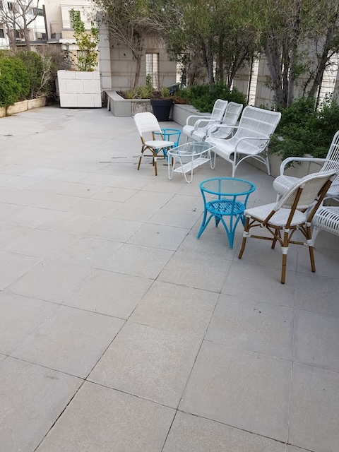 מרפסת חיצונית על גג במרכז תל אביב עם כיסאות קש לבנים, שולחן קפה מזכוכית, שולחנות צד כחולים וצמחים לאורך הקירות.