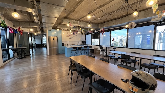 אזור מנוחה מרווח במשרד ברחוב יגאל אלון עם שולחנות ארוכים, כיסאות, דלפק מטבח, קישוטים חגיגיים, חלונות גדולים, מרפסת סמוכה וקסדת אופנוע על השולחן.