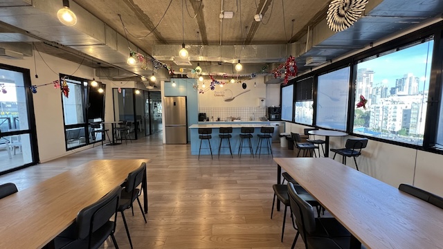 מטבח ומרחב אוכל מודרניים במשרד עם רצפות עץ, שולחנות, כיסאות, כסאות בר, חלונות גדולים, קישוטים חגיגיים, נוף לעיר ברקע ומרפסת המשקיפה על יגאל אלון.