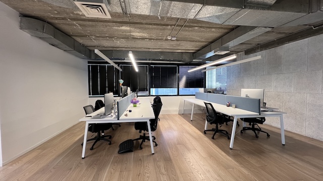 חלל משרדי מודרני עם ארבעה שולחנות, מחשבים וכסאות בשתי שורות, עם חלונות גדולים עם תריסים שחורים, ריצוף עץ, מרפסת המשקיפה על יגאל אלון וצינורות תקרה חשופים.