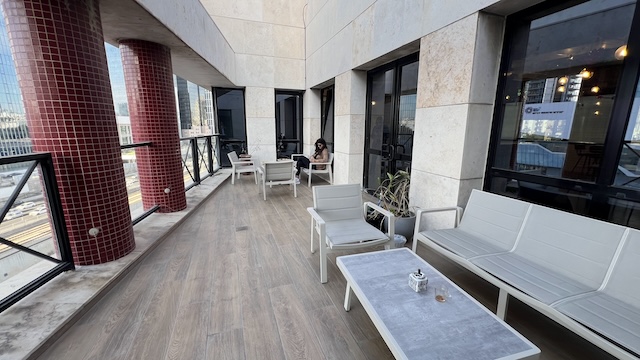 מרפסת חיצונית עם ספות ושולחנות בהירים לצד בניין עם חלונות גדולים ברחוב יגאל אלון; אדם יושב לבדו ליד שולחן ברקע.