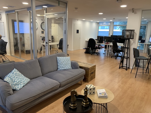 משרד מודרני להשכרה ברחוב החשמונאים, תל אביב, הכולל חדרי ישיבות עם קירות זכוכית, שולחנות עם מחשבים, ספה אפורה ושולחן קטן עם כלי שחמט וספרים על רצפת עץ.