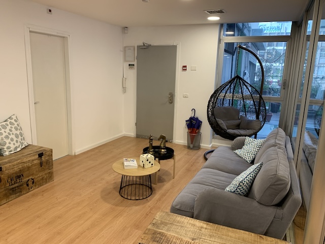 חדר המתנה מודרני בתל אביב עם ספה אפורה, כיסא תלוי, שולחן עגול קטן, ארגזי עץ וחלונות מהרצפה עד התקרה המכניסים אור טבעי — מושלם לכל משרד להשכרה.