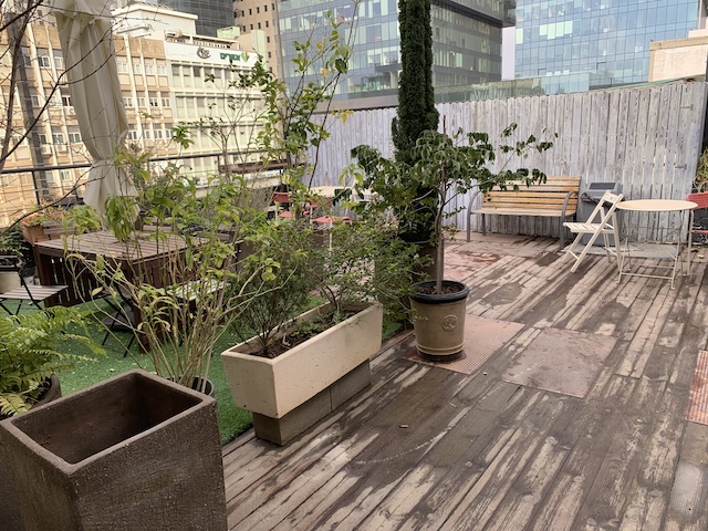 מרפסת גג מעץ עם עציצים, ספסלים ושולחנות, מוקפת גדר עץ ובניינים גבוהים — משרד עם מרפסת מושלם בחשמונאים תל אביב.