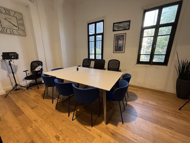 חדר ישיבות למשרד להשכרה ברחוב הרצל, תל אביב, הכולל שולחן מלבני גדול, שמונה כיסאות כחולים, שלושה כיסאות שחורים, חלונות, תמונות על הקירות, שעון, תאורת וידאו וצמח על רצפת עץ.