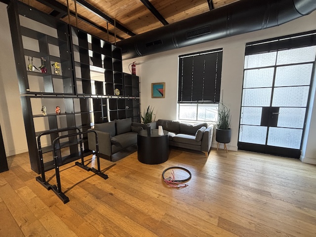 טרקלין משרדי מודרני בתל אביב עם מדף שחור, שני כיסאות אפורים, שולחן עגול, עציצים ופס התנגדות עגול על רצפת עץ. חלונות גדולים ממלאים את המשרד להשכרה באור טבעי.