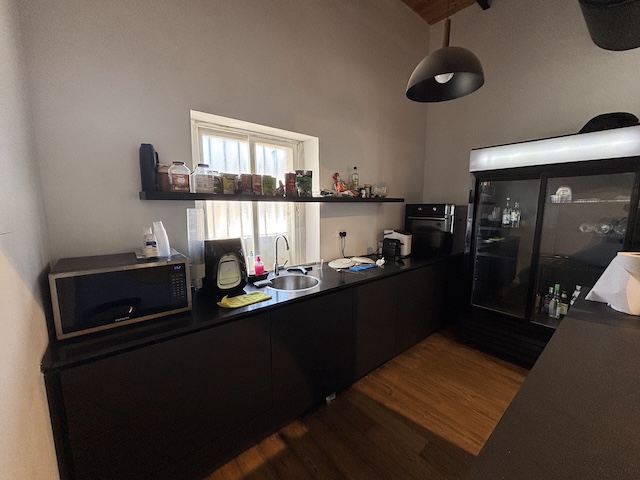 מטבח מודרני עם ארונות כהים, מכשירי חשמל על השיש, מיקרוגל, כיור ומקרר גדול — מושלם למשרד להשכרה בתל אביב. מדף מעל השיש מכיל פריטים שונים. רצפת עץ ותאורה תלויה מעל.