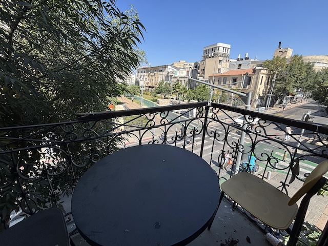 שולחן עגול שחור וכסא בודד ניצבים במרפסת המשקיפה על רחוב הרצל, עם בנייני העיר ועצים תחת שמי תל אביב הכחולים והצלולים.