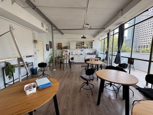 חלל עבודה מואר ומודרני עם שולחנות עץ עגולים, כיסאות, מטבחון בחלק האחורי וחלונות גדולים המשקיפים על נוף העיר — מושלם משרד להשכרה באיסרליש 6, תל אביב.