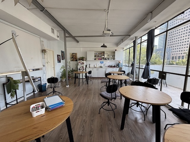 חלל עבודה מואר ומודרני עם שולחנות עץ עגולים, כיסאות, מטבחון בחלק האחורי וחלונות גדולים המשקיפים על נוף העיר — מושלם משרד להשכרה באיסרליש 6, תל אביב.