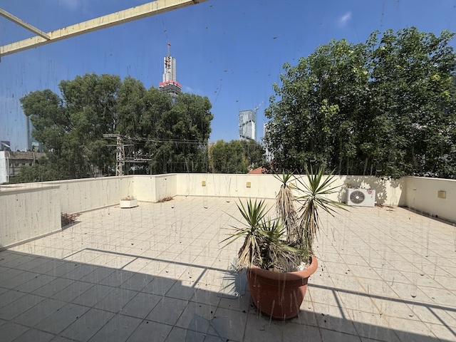 מרפסת גג מרוצפת עם עציץ גדול במרכזה, הנשקפת מבעד לחלון זכוכית מלוכלך באיזרליש 6, תל אביב — מושלמת למשרד להשכרה הבא שלכם, עם עצים עירוניים ובניינים גבוהים ברקע.