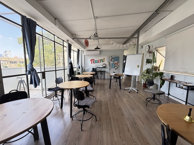 כיתה מוארת עם שולחנות עגולים, כיסאות, לוח לבן, צמחים, חלונות גדולים ושלט "Fluent" על הקיר האחורי — מושלמת לסדנאות או משרד להשכרה ליד מונטיפיורי תל אביב.