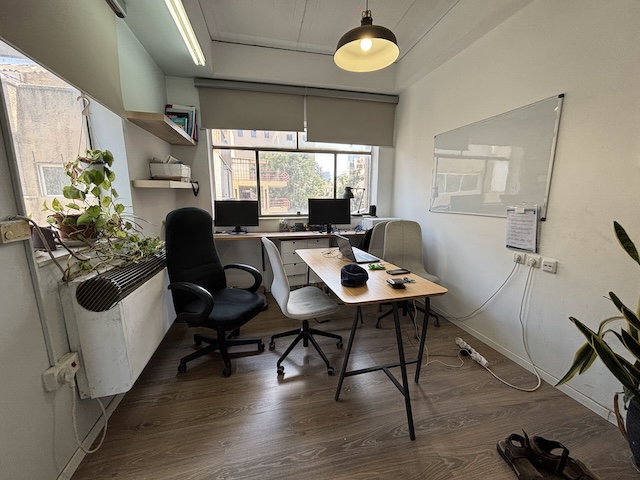 משרד קטן להשכרה עם שני שולחנות, כיסאות, מסכי מחשב, לוח לבן, עציצים וחלונות המאפשרים כניסת אור טבעי. ממוקם ברחוב איסרליש 6, תל אביב.