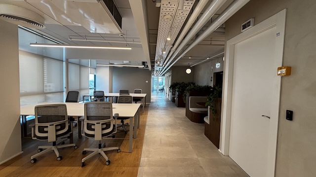 פנים משרד מודרני עם מספר שולחנות וכסאות ריקים, חלונות גדולים משמאל, ומסדרון עם צמחייה ודלתות סגורות מימין, מושלם עבור אלה המחפשים משרדים להשכרה בבית ישראכרט.