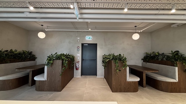 ארבעה תאי ישיבה מעץ עם כריות אפורות וצמחים ירוקים מקיפים את קירותיו של משרד מרוהט מודרני בגוונים ניטרליים בתל אביב; דלת שחורה ותאורת תקרה עגולה נראות לעין.