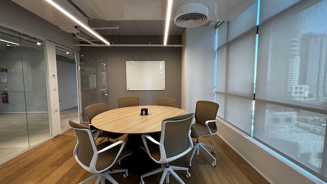 חדר ישיבות מודרני בבית ישראכרט כולל שולחן עץ עגול, שישה כיסאות, לוח לבן, חלונות גדולים עם תריסים ורצפת עץ — אידיאלי לעסקים המחפשים משרד מודרני להשכרה.