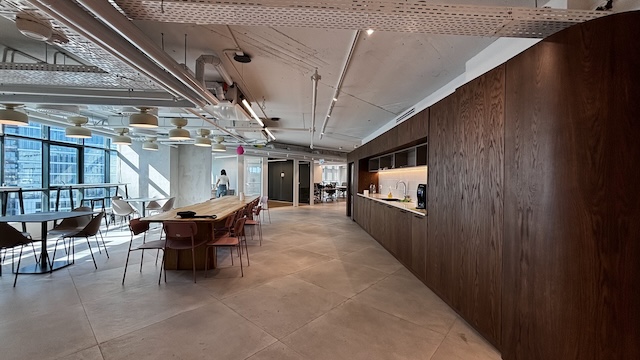 מטבח ומטבח מודרני עם שולחנות עץ, כיסאות, ארונות מובנים וחלונות גדולים המשקיפים על בנייני העיר בבית ישראכרט. אדם אחד עומד ליד החלונות במשרד מודרני להשכרה, 800 מ"ר.