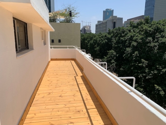 מרפסת עץ עם מעקות לבנים משקיפה על אזור מיוער ובניינים גבוהים בשדרות רוטשילד, ומציעה נוף עירוני וקסם שטוף שמש — מושלם למשרד להשכרה בלב העיר.