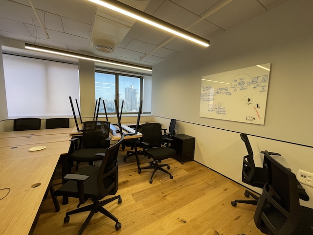 משרד ריק עם רצפת עץ, כיסאות מוערמים על שולחנות, חלון גדול המשקיף על בנייני העיר במתחם הבורסה, ולוח לבן עם הערות על הקיר — משרד גדול להשכרה בבית אבגד.