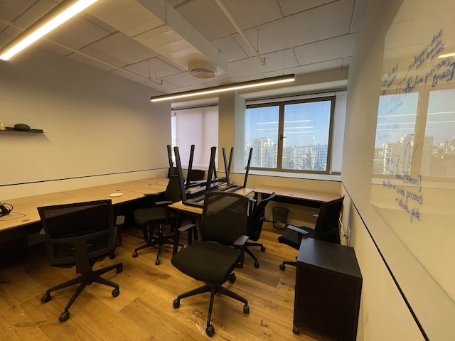 משרד ריק עם רצפת עץ בבית אבגד, מספר שולחנות וכסאות מוערמים, חלון עם נוף לעיר מתחם הבורסה, ולוח לבן עם כיתוב על הקיר הימני — משרד גדול להשכרה.