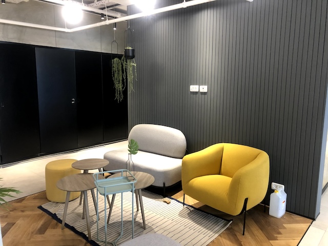 פינת ישיבה מודרנית עם כורסה צהובה, ספה זוגית אפורה, שולחנות קפה מקוננים, שטיח פסים וצמח תלוי על רקע קיר מחופה לוחות כהים — מושלמת למשרד להשכרה הבא שלכם במתחם הבורסה.