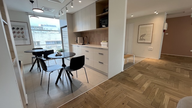 מטבח מודרני ופתוח ופינת אוכל עם כיסאות שחורים, שולחנות עגולים, ארונות מעץ בהיר וריצוף פרקט, הצמוד למסדרון עם יצירות אמנות — מושלם למשרד להשכרה בבית ג'ימיני בתל אביב.