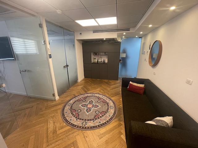 אזור המתנה מודרני במשרד בתל אביב עם ספה כהה, שטיח עגול עם דוגמה, חדר עם קירות זכוכית ומראה המותקנת על הקיר. כולל ריצוף עץ בדוגמת אריחים, תאורה תקרתית וגישה למרפסת. משרד מושלם להשכרה.