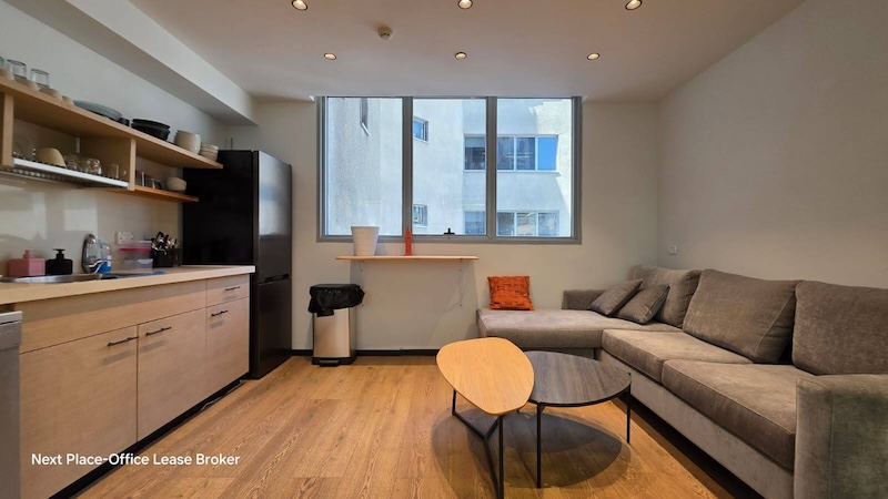 אזור ישיבה מודרני במשרד עם ספה פינתית אפורה, שולחן קפה עגול, ריצוף עץ ומדפי מטבח פתוחים – מושלם למשרד להשכרה במנחם בגין, תל אביב. חלונות גדולים ותאורה תקרתית יוצרים סביבת עבודה מזמינה.