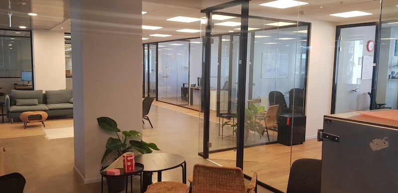 משרד מרוהט ומודרני עם חדרי ישיבות בעלי קירות זכוכית, שולחנות, כיסאות, צמחים מקורה ואזור ישיבה עם ספה ושולחן קפה. ממוקם ברחוב יגאל אלון בתל אביב, עם תאורה בהירה ועיצוב נקי ומסודר.