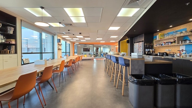 מטבח משרדי מודרני ופינת ישיבה עם שולחנות ארוכים, כיסאות צבעוניים, שרפרפים גבוהים וחלונות גדולים המכניסים אור טבעי – אידיאלי למשרד להשכרה במתחם הבורסה רמת גן.