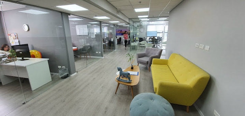 משרד להשכרה בתל אביב, עם חדרים עם קירות זכוכית, ספה צהובה, שולחן קטן, ריצוף עץ בהיר ומרפסת; שולחנות וכסאות מסודרים ברקע.