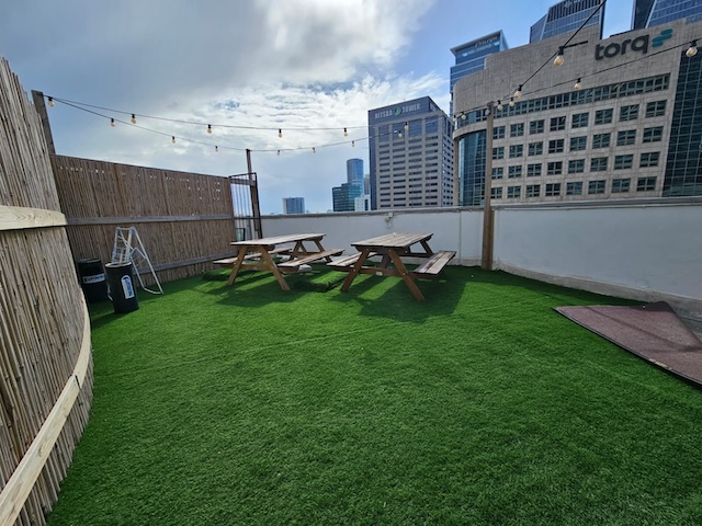 אזור גג פלאג&פליי, מרוהט במלואו בדשא מלאכותי, שולחנות פיקניק מעץ, אורות מחרוזת, ונוף של בנייני העיר ליד מנחם בגין שרונה תחת שמיים מעוננים חלקית.