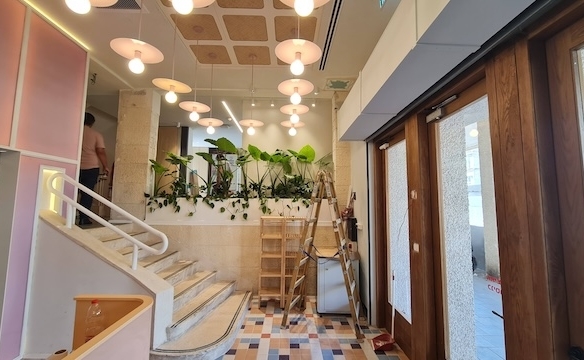 פנים מודרני בבניין להשכרה עם דלתות עץ, רצפת אריחים, גרם מדרגות, מנורות תלייה ועציץ עם צמחים ירוקים. סולם וחומרי ניקוי נמצאים ליד הקיר.