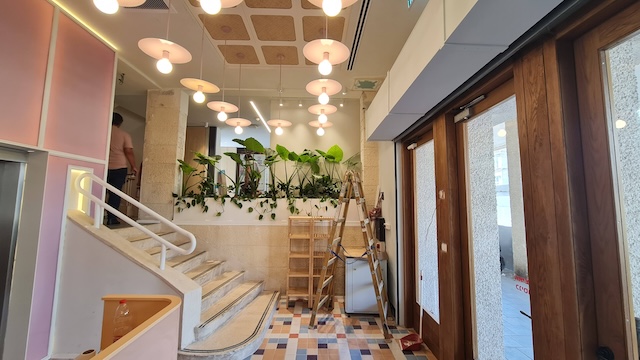 פנים מודרני בבניין להשכרה עם דלתות עץ, רצפת אריחים, גרם מדרגות, מנורות תלייה ועציץ עם צמחים ירוקים. סולם וחומרי ניקוי נמצאים ליד הקיר.