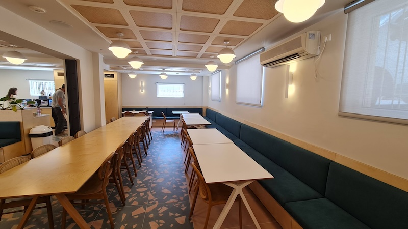 פינת אוכל מודרנית עם שולחנות ארוכים, כיסאות עץ, ספסלי ישיבה מרופדים בירוק, רצפת אריחים ותאורת תקרה בבניין עצמאי ברחוב אבן גבירול בתל אביב. כמה אנשים נראים עומדים ליד הכניסה. מושלם להשכרה.