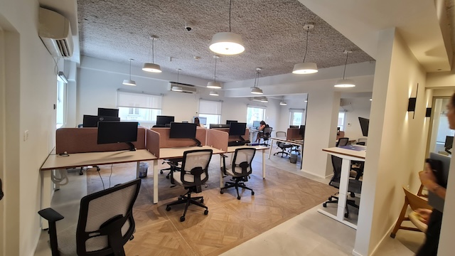 חלל משרדים מודרני ופתוח עם כיסאות ארגונומיים, שולחנות ריקים ומסכי מחשב להשכרה בבניין עצמאי באבן גבירו, תל אביב; כמה אנשים עובדים בקצה הרחוק תחת תאורה חזקה.