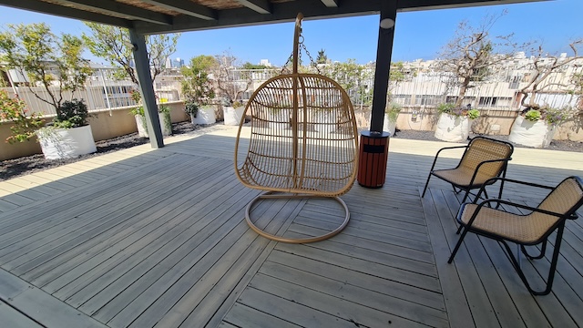 מרפסת עץ עם כיסא נצרים תלוי, שני כיסאות תואמים, פח אשפה ועציצים מתחת לפרגולה בבניין עצמאי באבן גבירול בתל אביב, עם נוף עירוני הנשקף ברקע — מושלם להשכרה.