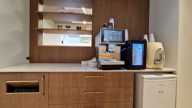 מטבחון מעץ עם מדפים, מכונת קפה, מקרר קטן, כוסות מוערמות ומגבות נייר על משטח עבודה לבן — מושלם להשכרה בבניין עצמאי ליד אבן גבירול בתל אביב.