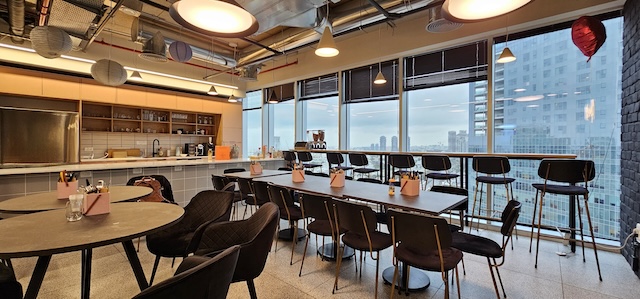 מטבח משרדי מודרני ופינת אוכל עם שולחנות, כיסאות, מקומות ישיבה ליד חלונות גדולים ונוף מרהיב של קו הרקיע של תל אביב ועזריאלי טאון. משרד להשכרה מושלם לצוותים המחפשים סגנון ונוחות.