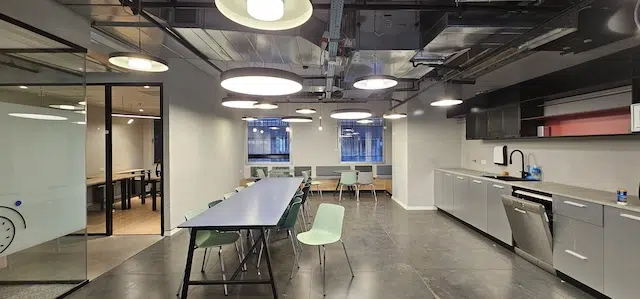 מטבח משרדי מודרני עם שולחן ארוך, כיסאות ירוקים, מכשירי חשמל מובנים, תאורה תקרתית וחדר ישיבות עם קירות זכוכית ברקע — Plug & Play משרד להשכרה ב-עמינדב תל אביב.