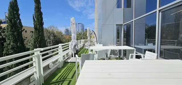 אזור פטיו חיצוני עם שולחנות וכסאות לבנים, ריצוף דשא מלאכותי ושמשיות, הצמוד לבניין זכוכית מודרני תחת שמיים כחולים וצלולים — מושלם עבור משרד להשכרה Plug & Play בעמינדב תל אביב.