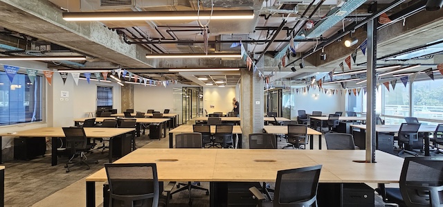 משרד להשכרה בעמינדב תל אביב, בעל חלל פתוח גדול, עם שורות של שולחנות וכסאות ריקים, תקרות חשופות, קירות זכוכית ודגלים צבעוניים – חלל עבודה מסוגנן מסוג Plug & Play המוכן לקלוט את הצוות שלכם.