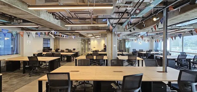 משרד להשכרה בעמינדב תל אביב, בעל חלל פתוח גדול, עם שורות של שולחנות וכסאות ריקים, תקרות חשופות, קירות זכוכית ודגלים צבעוניים – חלל עבודה מסוגנן מסוג Plug & Play המוכן לקלוט את הצוות שלכם.