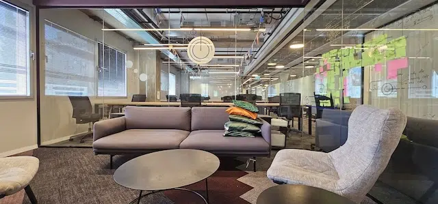 טרקלין משרדי מודרני במשרד להשכרה עמינדב תל אביב, הכולל ספה אפורה, כורסה, שולחנות שחורים, קירות זכוכית ופתקיות צבעוניות על לוח — חלל עבודה מושלם מסוג Plug & Play.