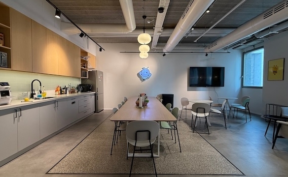 מטבח משרדי מודרני ופינת ישיבה להשכרה במגדל מילניום, הכולל שולחן ארוך, כיסאות, מטבחון משמאל, ספות מימין וטלוויזיה גדולה התלויה על הקיר. אור טבעי שוטף את החדר מבעד לחלונות.