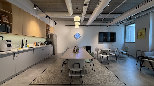 מטבח משרדי מודרני ופינת ישיבה להשכרה במגדל מילניום, הכולל שולחן ארוך, כיסאות, מטבחון משמאל, ספות מימין וטלוויזיה גדולה התלויה על הקיר. אור טבעי שוטף את החדר מבעד לחלונות.