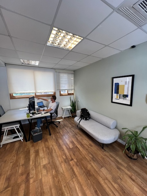 אדם יושב ליד שולחן עבודה ומחשב במשרד להשכרה מודרני בתל אביב, עם רצפות עץ, צמחים, ספה אפורה ואמנות קיר מופשטת.