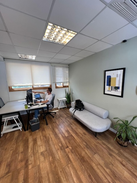 אדם עובד ליד שולחן עם שני מסכים במשרד להשכרה, 80 מ"ר, ביוסף קארו תל אביב, עם רצפות עץ, ספה אפורה, עציצים ויצירות אמנות מופשטות ממוסגרות על הקיר.