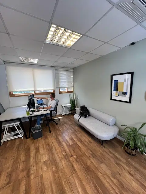 אדם עובד ליד שולחן עם שני מסכים במשרד להשכרה, 80 מ"ר, ביוסף קארו תל אביב, עם רצפות עץ, ספה אפורה, עציצים ויצירות אמנות מופשטות ממוסגרות על הקיר.