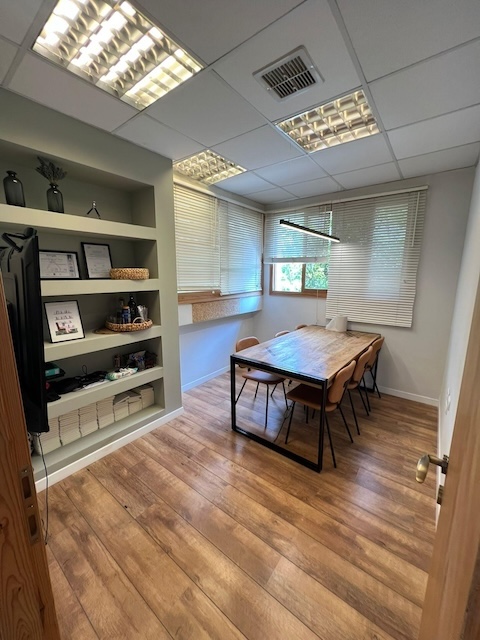 משרד קטן להשכרה ברחוב יוסף קארו בתל אביב, הכולל שולחן עץ, שישה כיסאות, מדפים מובנים, טלוויזיה וחלונות גדולים עם תריסים המאפשרים כניסת אור טבעי.